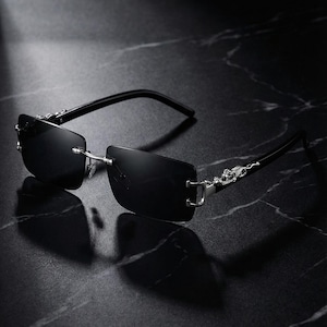 May include: Black rimless sunglasses with rectangular lenses. The temples are black with silver decorative accents. The sunglasses are resting on a dark, marble-like surface.
