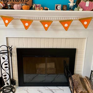 May include: A handmade crochet pie slice banner with orange and yellow colors. The banner is strung across a white fireplace mantel, with a black fireplace below. Decorative items are on the mantel.