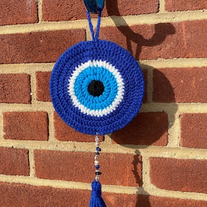 May include: A blue crochet evil eye wall hanging with a black centre, surrounded by white and light blue rings. A blue tassel and beaded strand hang below. The wall hanging is suspended from a blue hook against a red brick wall.