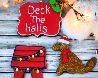 May include: Christmas-themed cookies. One cookie is a red sign with the text "Deck The Halls". Another cookie is a red dog house with colorful lights. The third cookie is a dog wearing a Santa hat and red scarf.