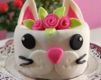 May include: A white fondant cake decorated with pink and white frosting to look like a cat. The cake has pink ears, a pink nose, and black eyes. The cake is decorated with pink and white flowers.