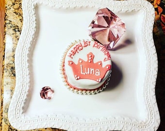 May include: A white cake with pink frosting and a pink crown with the name "Luna" on it. The cake is decorated with white pearls and is on a white plate with a pink diamond.