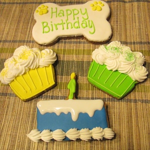 Pode incluir: Três bolachas com tema de cão: uma bolacha em forma de osso com o texto "Happy Birthday", um cupcake amarelo com cobertura branca e um cupcake verde com cobertura branca.