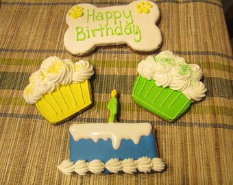 May include: Three dog-themed cookies: a bone-shaped cookie with the text "Happy Birthday", a yellow cupcake with white frosting, and a green cupcake with white frosting.