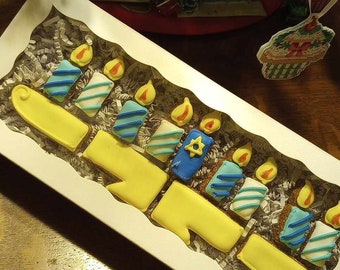 May include: A box of Hanukkah-themed cookies shaped like a menorah. The cookies are yellow, blue, and white, with flame-shaped icing. The menorah cookies are arranged in a white box with shredded paper, set on a wooden table.