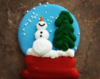May include: A round cookie decorated with blue icing to resemble a snow globe. It features a white snowman with an orange nose, a green pine tree, and white icing dots representing falling snow, all set on a solid red base.