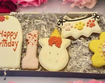 May include: A set of five dog birthday cookies in a box. The cookies are decorated with pink, yellow, and white frosting. The cookies are shaped like a heart, a number one, a unicorn, a banner, and a flower. The heart cookie says "Happy Birthday".