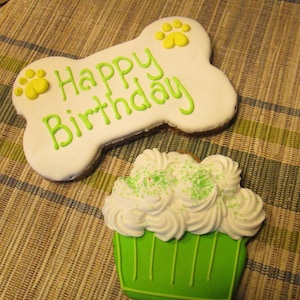 May include: A white bone-shaped dog treat with green icing and the words "Happy Birthday" and two yellow paw prints. A green cupcake with white frosting and green sprinkles is next to the bone.