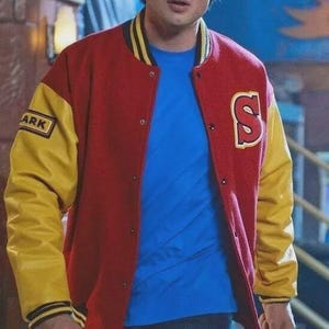 Op de afbeelding: Een rood met gele varsity-jas met een letter S-patch op de linkerborst en een ARK-patch op de rechtermouw. De jas heeft een drukknoopsluiting, een geribbelde kraag en boorden met gele strepen, gedragen over een blauw t-shirt en een spijkerbroek.
