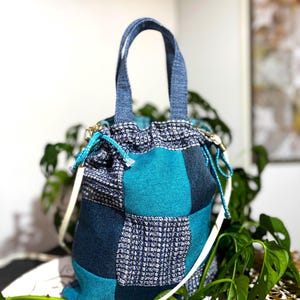 Può includere: Una borsa tote patchwork con chiusura a coulisse. La borsa presenta un mix di tessuti blu e turchese, con una tracolla bianca e manici blu. La borsa è alta circa 30 cm.