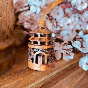 May include: A small, intricately carved wooden pagoda ornament with a gold ribbon for hanging. The pagoda is brown with detailed cutouts and sits on a wooden surface, with white and light blue flowers in the background.