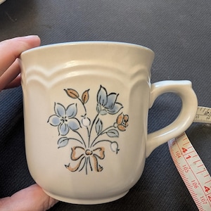 Puede incluir: Taza de cerámica color crema con diseño floral. La taza presenta un ramo de flores azules y naranjas atado con un lazo marrón. El asa es cuadrada y el borde tiene un borde festoneado. Una cinta métrica está al lado de la taza.