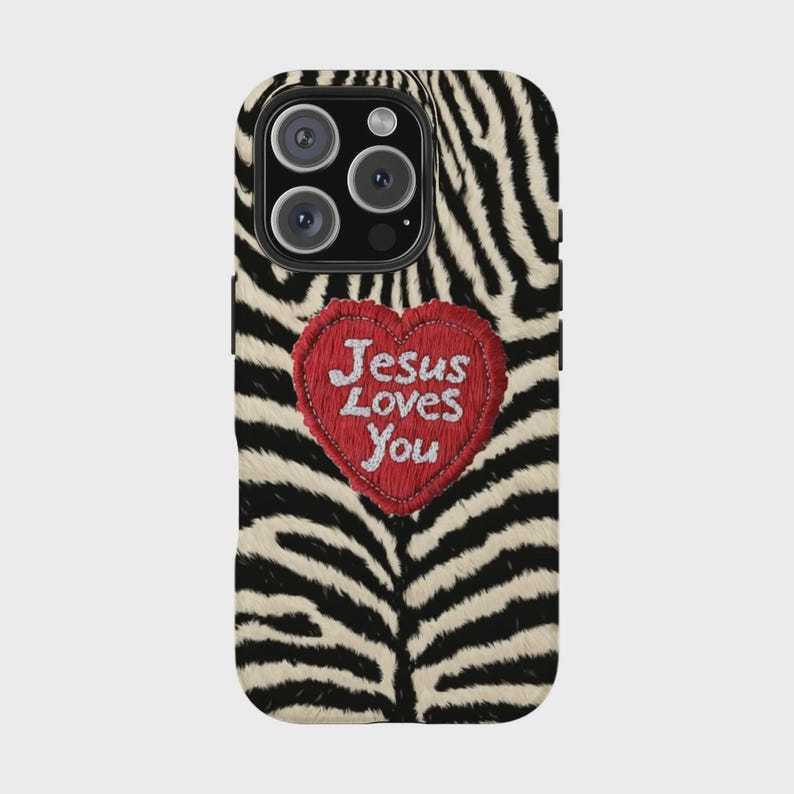 K&ouml;nnte beinhalten: Eine Handyh&uuml;lle mit Zebramuster und einem herzf&ouml;rmigen roten Aufn&auml;her. Der Aufn&auml;her hat den gestickten Text "Jesus Loves You" in Wei&szlig;. Die H&uuml;lle hat ein schwarzes Kameramodul.