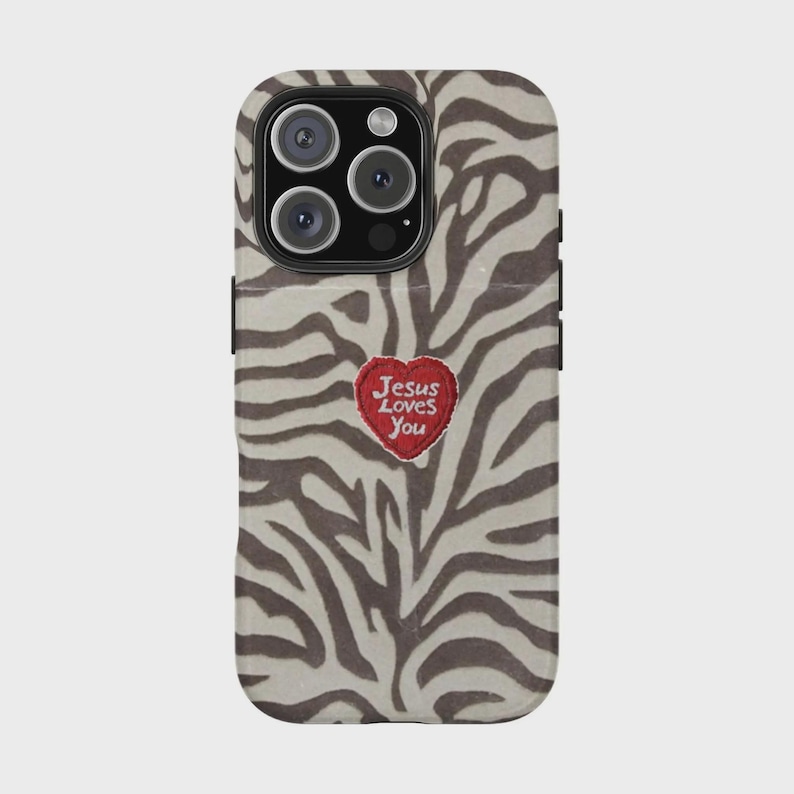 K&ouml;nnte beinhalten: Eine Handyh&uuml;lle mit einem Zebra-Print-Design in Braun- und Beiget&ouml;nen. Ein roter herzf&ouml;rmiger Aufn&auml;her mit dem Text "Jesus Loves You" ist auf der H&uuml;lle zentriert. Die H&uuml;lle hat einen Kameraausschnitt und schwarze R&auml;nder.
