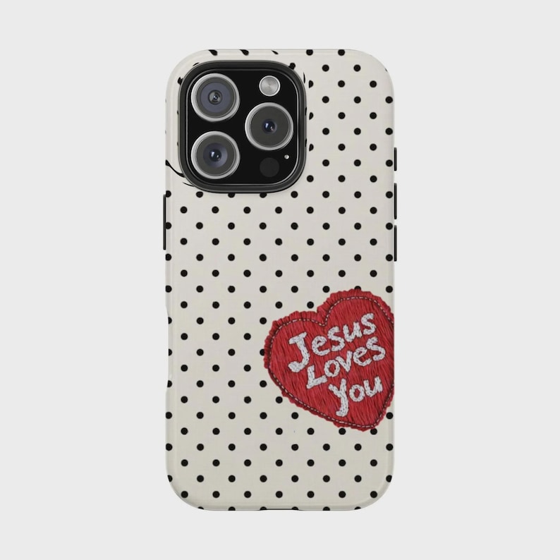 K&ouml;nnte beinhalten: Eine Handyh&uuml;lle mit wei&szlig;em Hintergrund und schwarzen Punkten. Ein rotes Herz mit dem Text "Jesus Loves You" ist auf der H&uuml;lle gestickt. Die Handyh&uuml;lle hat eine Kamera und einen Blitz.