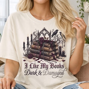 May include: A cream-coloured t-shirt with a graphic design featuring a stack of books, candles, roses, and ravens. The text "I Like My Books Dark & Damaged" is displayed below the graphic. The shirt is made of a soft material.