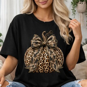 Op de afbeelding: Een zwart t-shirt met korte mouwen en een grote afbeelding van een pompoen met een luipaardprint en een bijpassende strik om de steel. Het shirt wordt gedragen met een lichtblauwe spijkerbroek met gescheurde gaten bij de knieën.