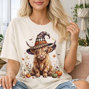 May include: A cream-colored t-shirt featuring a graphic of a fluffy Highland cow wearing a witch hat with a checkered pattern. The design includes small pumpkins, autumn leaves, crescent moons, and tiny ghosts surrounding the cow.