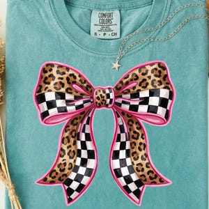 May include: A teal t-shirt with a large bow design. The bow features a leopard print pattern with a pink outline and checkered accents. A silver necklace with a star pendant is draped on the shirt.