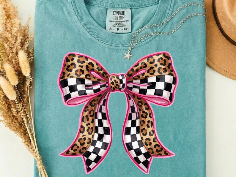 May include: A teal t-shirt with a large bow design. The bow features a leopard print pattern with a pink outline and checkered accents. A silver necklace with a star pendant is draped on the shirt.