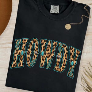 以下が含まれることがあります： ヒョウ柄のデザインで「HOWDY」という文字がプリントされた黒いTシャツ。 ターコイズとシルバーのアクセントが施されています。 シャツはクルーネックで半袖です。 ペンダント付きのゴールドネックレスも確認できます。