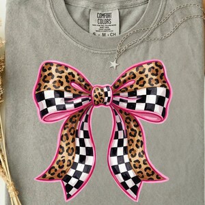 May include: A grey t-shirt with a large bow design. The bow has a pink outline, leopard print sections, and black and white chequered sections. A silver necklace with a star pendant and a hat are also visible.
