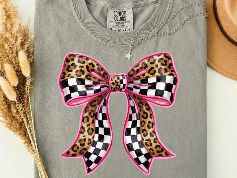 May include: A grey t-shirt with a large bow design. The bow has a pink outline, leopard print sections, and black and white chequered sections. A silver necklace with a star pendant and a hat are also visible.