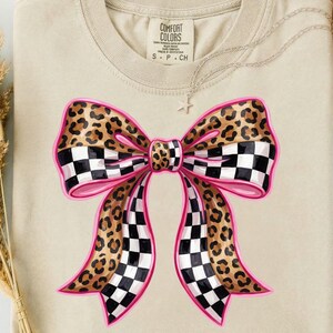 May include: A beige Comfort Colors t-shirt with a large bow design. The bow has a pink outline, leopard print sections, and black and white checkered sections. A silver necklace with a star pendant is visible.