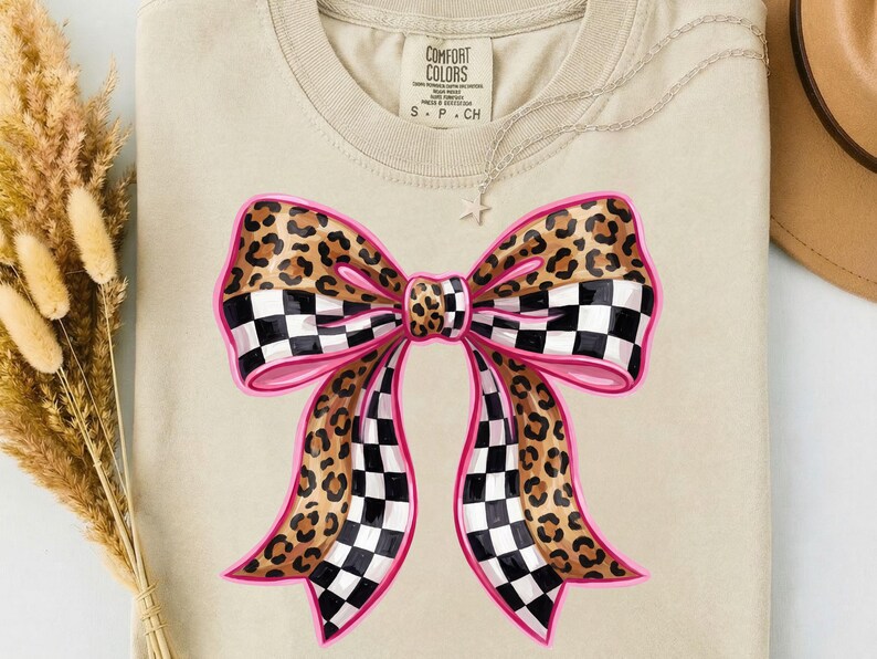 May include: A beige Comfort Colors t-shirt with a large bow design. The bow has a pink outline, leopard print sections, and black and white checkered sections. A silver necklace with a star pendant is visible.