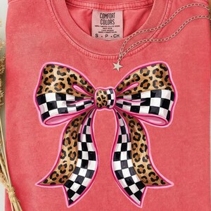 May include: A coral-coloured t-shirt with a large bow design. The bow has a pink outline, a checkered pattern, and a leopard print. A silver necklace with a star pendant is draped across the shirt. The tag reads "COMFORT COLORS".