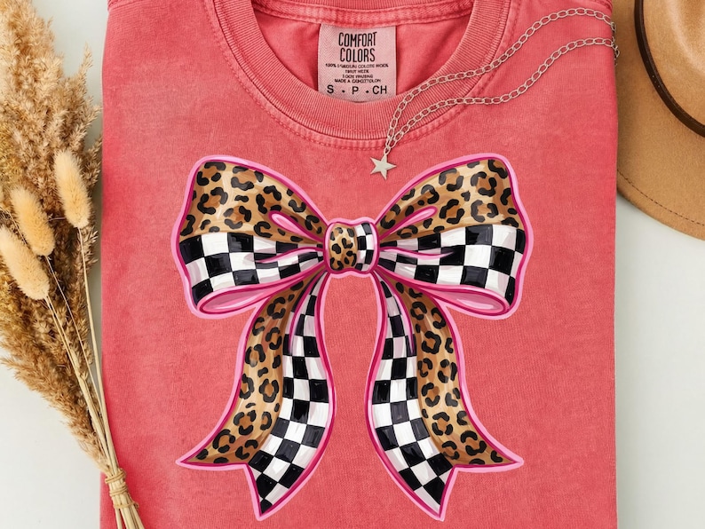 May include: A coral-coloured t-shirt with a large bow design. The bow has a pink outline, a checkered pattern, and a leopard print. A silver necklace with a star pendant is draped across the shirt. The tag reads "COMFORT COLORS".