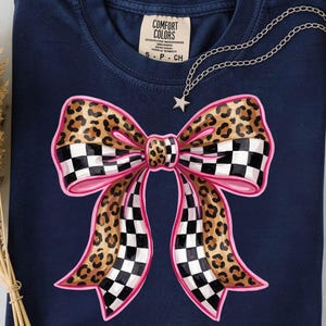 May include: Navy blue t-shirt with a large bow design. The bow features a leopard print centre, pink outline, and a checkered pattern on the ends. A silver necklace with a star pendant is draped on the shirt.