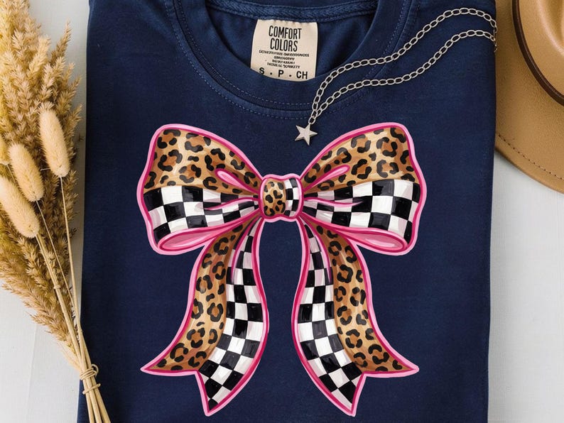 May include: Navy blue t-shirt with a large bow design. The bow features a leopard print centre, pink outline, and a checkered pattern on the ends. A silver necklace with a star pendant is draped on the shirt.