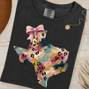 Puede incluir: Camiseta gris oscuro con un diseño colorido en forma de Texas y un lazo rosa. El diseño incorpora pinceladas de color turquesa, rosa, dorado y amarillo con manchas de leopardo negras. Se ve un collar dorado y un sombrero marrón.