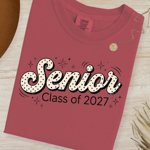 Puede incluir: Una camiseta granate con la palabra "Senior" en una fuente retro con lunares negros y un contorno negro. Debajo está el texto "Class of 2027". La camiseta tiene cuello redondo y mangas cortas. La camiseta es de la marca Comfort Colors.