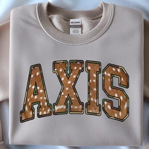 May include: A beige sweatshirt with the word "AXIS" printed across the front. The letters are filled with a brown and white spotted pattern, outlined in green and brown. The sweatshirt has a crew neck and long sleeves.