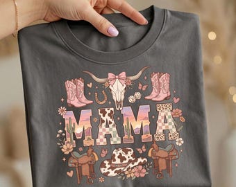 May include: Dark gray t-shirt featuring a colorful graphic design with the word "MAMA" in a Western-themed font. The design includes a cow skull, cowboy boots, a horseshoe, a cowboy hat, and other Western-themed elements.