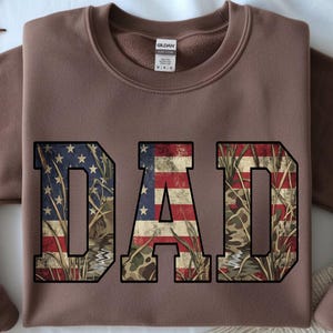 May include: A brown sweatshirt with the word "DAD" in large, distressed letters. The letters are filled with a combination of the American flag and a camouflage pattern. The sweatshirt has a crew neck and long sleeves.