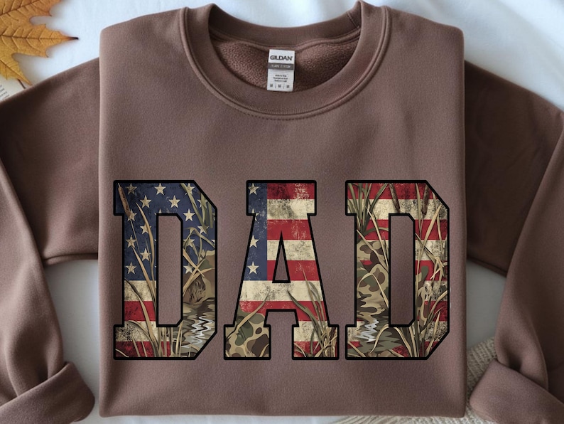 May include: A brown sweatshirt with the word "DAD" in large, distressed letters. The letters are filled with a combination of the American flag and a camouflage pattern. The sweatshirt has a crew neck and long sleeves.