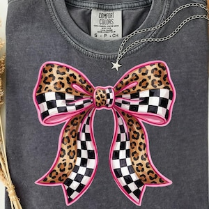 May include: Charcoal grey t-shirt with a large bow graphic. The bow features a pink outline, a black and white checkered pattern, and a leopard print design. A silver necklace with a star pendant is on the shirt. The tag reads "COMFORT COLORS".