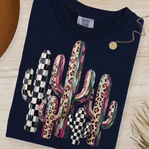 May include: Navy blue t-shirt featuring a colorful cactus design. The cacti have a mix of patterns, including checkered and leopard print, with pink, green, and brown accents. The shirt has a crew neck and the tag reads "Comfort Colors".