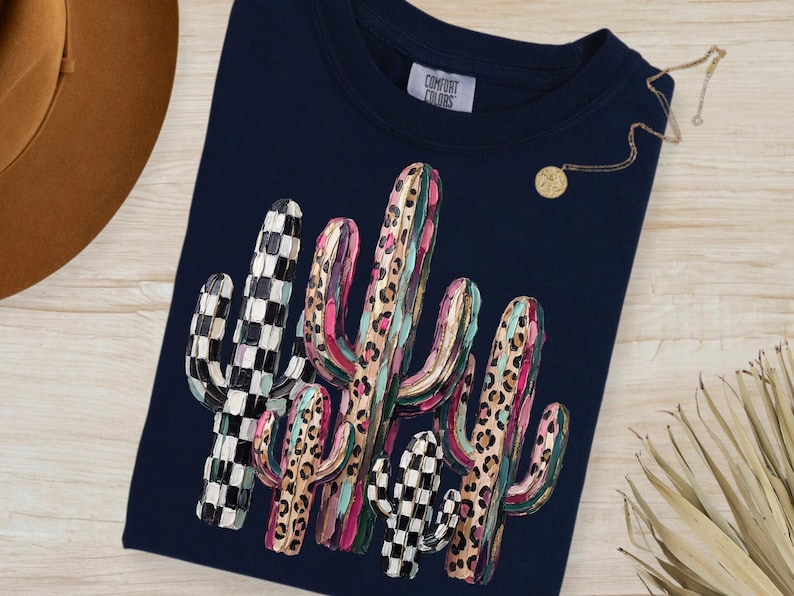 May include: Navy blue t-shirt featuring a colorful cactus design. The cacti have a mix of patterns, including checkered and leopard print, with pink, green, and brown accents. The shirt has a crew neck and the tag reads "Comfort Colors".