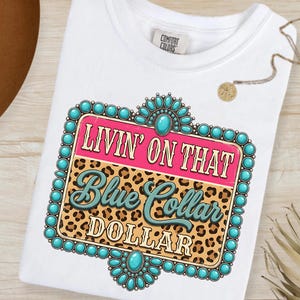 May include: White t-shirt with a graphic design. The design features the text "LIVIN' ON THAT Blue Collar DOLLAR" in a turquoise and pink color scheme, set against a leopard print background and turquoise beaded frame.
