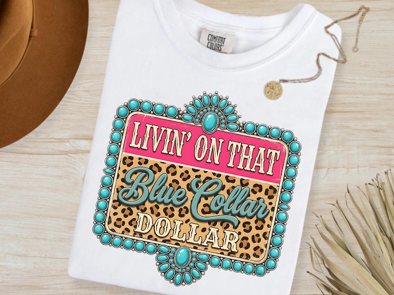 May include: White t-shirt with a graphic design. The design features the text "LIVIN' ON THAT Blue Collar DOLLAR" in a turquoise and pink color scheme, set against a leopard print background and turquoise beaded frame.