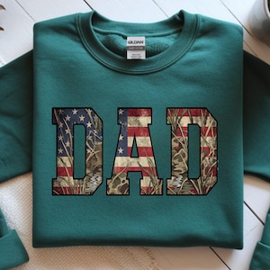 May include: Teal sweatshirt with the word "DAD" in large, bold letters. The letters are filled with a design that combines the American flag with a camouflage pattern. The sweatshirt is made of a soft material and has a crew neck.