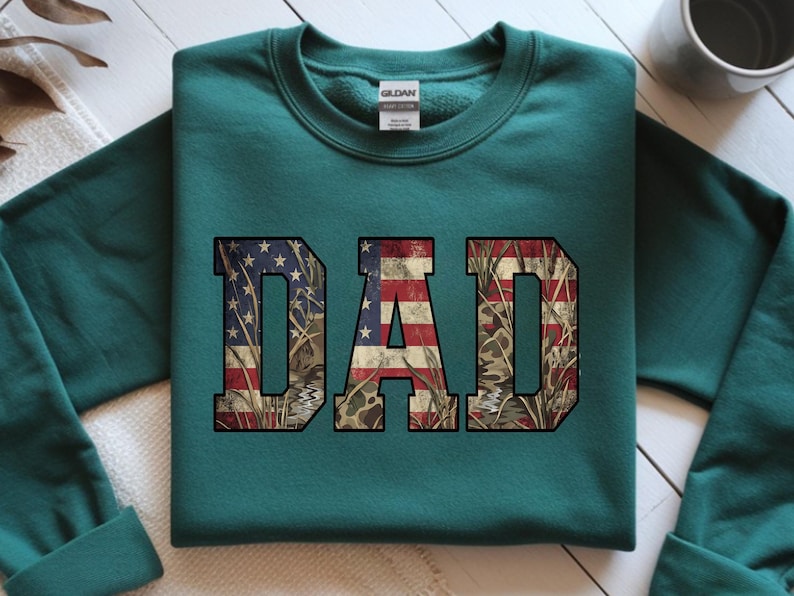 May include: Teal sweatshirt with the word "DAD" in large, bold letters. The letters are filled with a design that combines the American flag with a camouflage pattern. The sweatshirt is made of a soft material and has a crew neck.