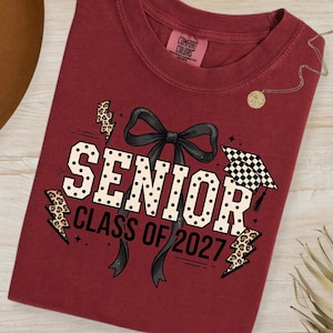 以下が含まれることがあります： 卒業デザインのマルーン色のComfort Colors Tシャツ。「SENIOR」の文字は白い水玉模様、黒いリボン、「CLASS OF 2027」の文字が入っています。ヒョウ柄の稲妻と卒業キャップがデザインを完成させています。