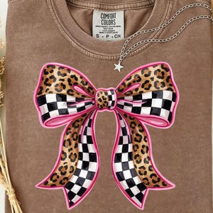 May include: A brown Comfort Colors t-shirt with a large bow design. The bow features a leopard print, a black and white checkered pattern, and pink outlines. A silver necklace with a star pendant is draped across the shirt.
