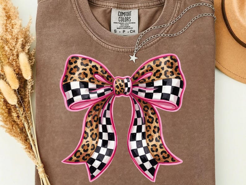 May include: A brown Comfort Colors t-shirt with a large bow design. The bow features a leopard print, a black and white checkered pattern, and pink outlines. A silver necklace with a star pendant is draped across the shirt.