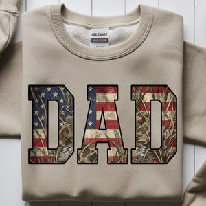 May include: A tan sweatshirt with the word "DAD" in large, block letters. The letters are filled with a distressed American flag design and camouflage pattern. The sweatshirt has a crew neck and long sleeves.
